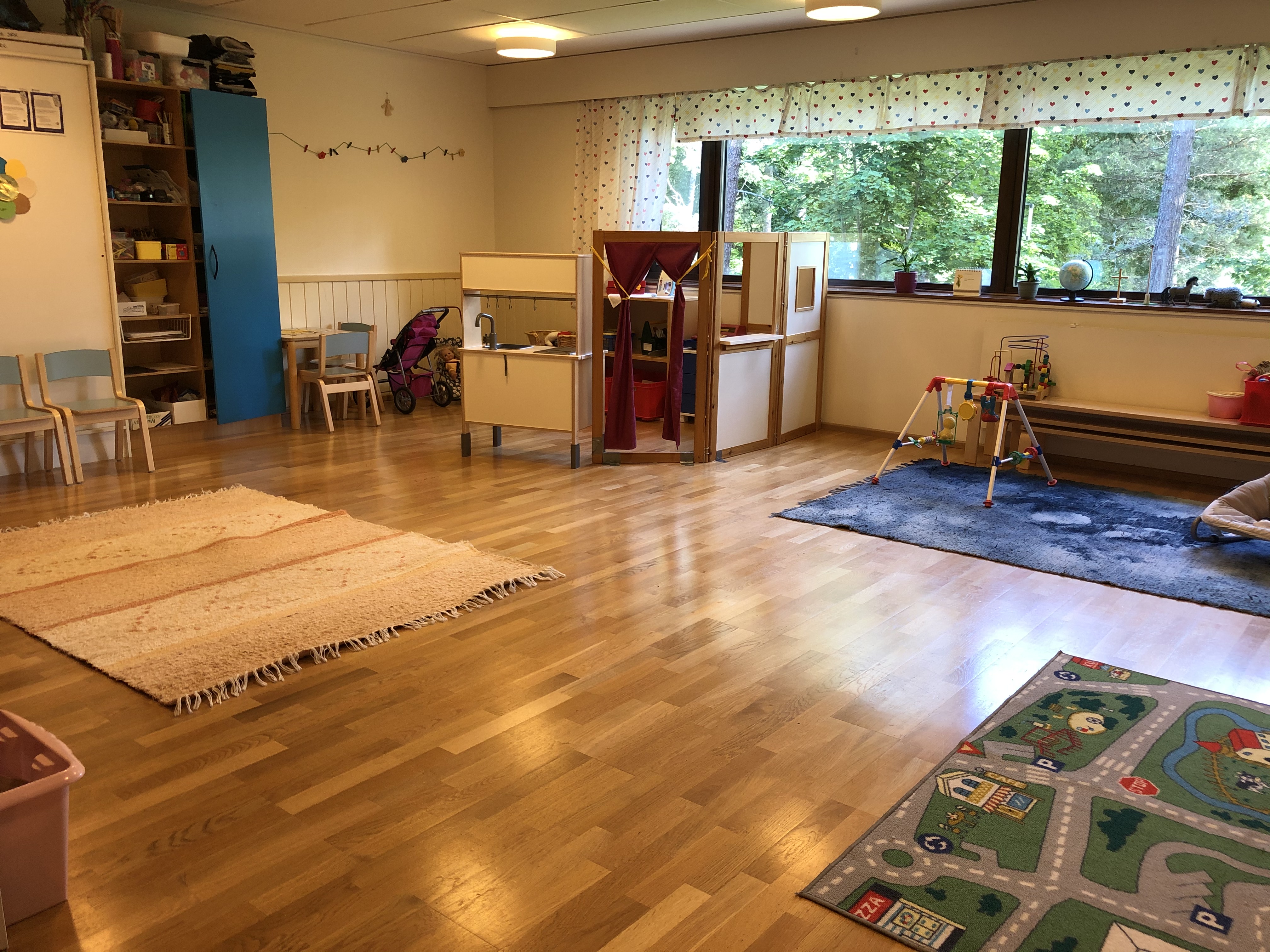 Perhekahvila Drop-in Kuva kerho huoneesta missä on etualalla leikkimatto autoille ja taustalla isot ikkunat ja keittiö leikki nurkka ja vauvalle leikki tilaa.