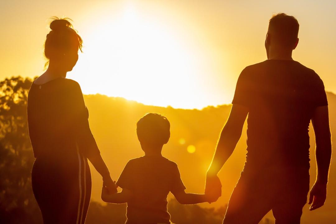 Nainen ja mies kävelevät auringon laskussa ja pitävät kädestä lasta, joka on heidän välissä