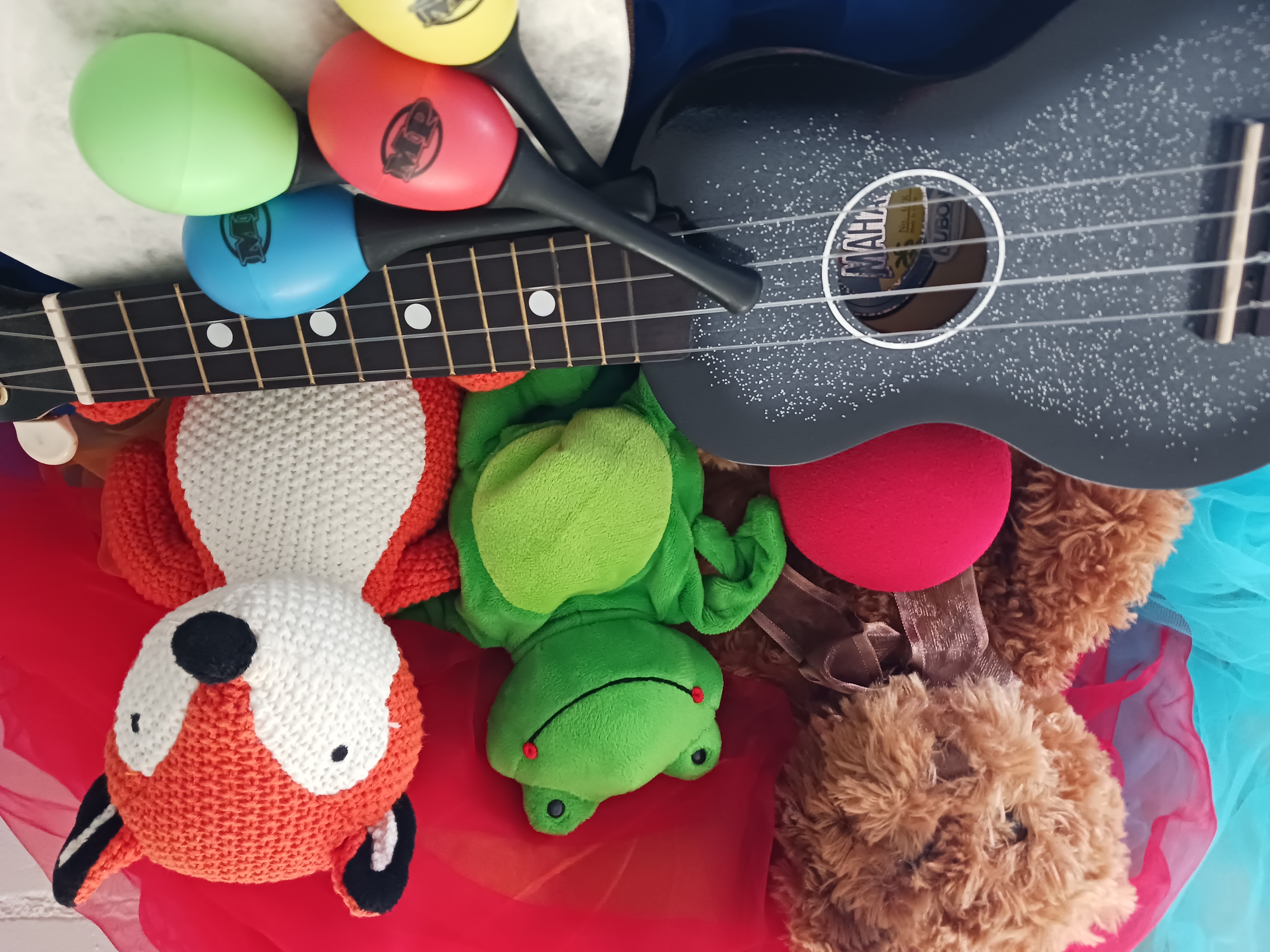Kuvassa on harmaa ukulele, värikkäitä marakaseja ja pehmoleluja.